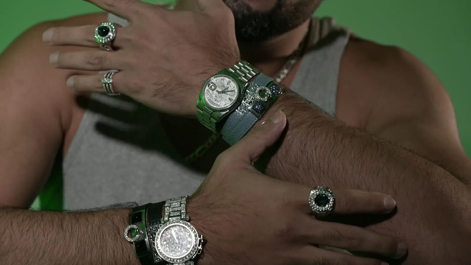 Crossed arms wearing Mozafarian rings, watches and bracelets against a green studio backdrop